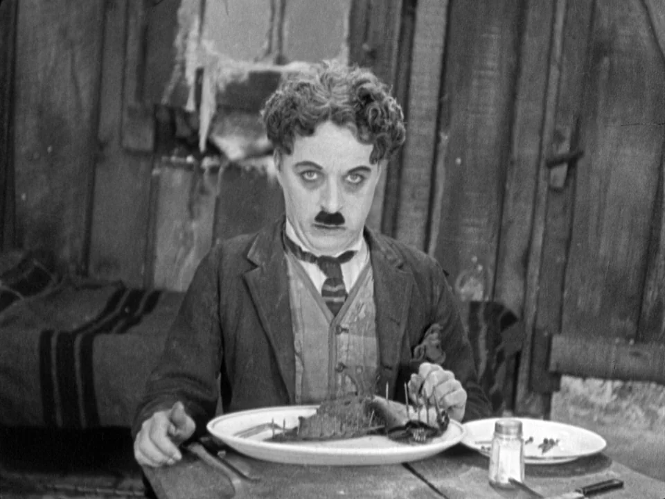 The Gold Rush (1925) movie still: medium — A rustic shack with weathered wooden wall planks serves as the backdrop. A man sits at a table and…