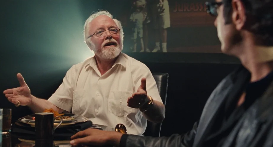 Jurassic Park (1993) movie still: medium — Two men sit at a table in a dimly lit setting. An older man with glasses gesticulates while talking.