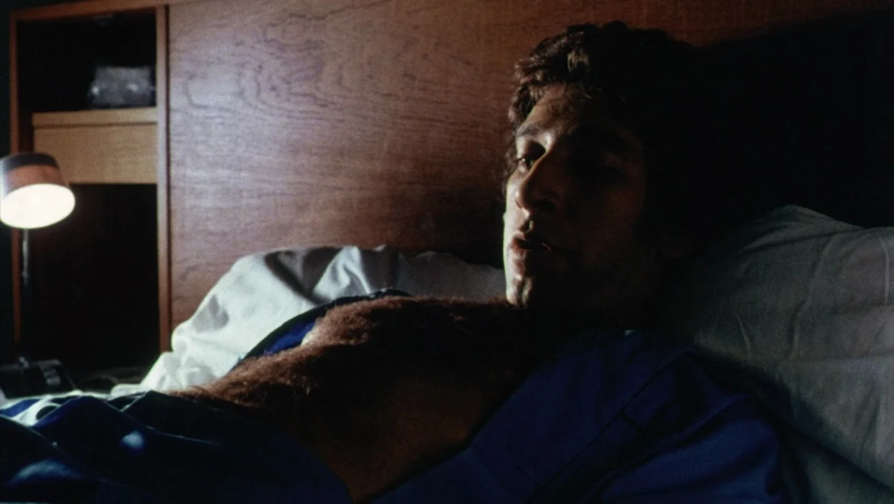 Shivers (1975) movie still: medium — A man lies in a bed against a wooden headboard in a dark room. The subject is partially illuminated by a lamp…