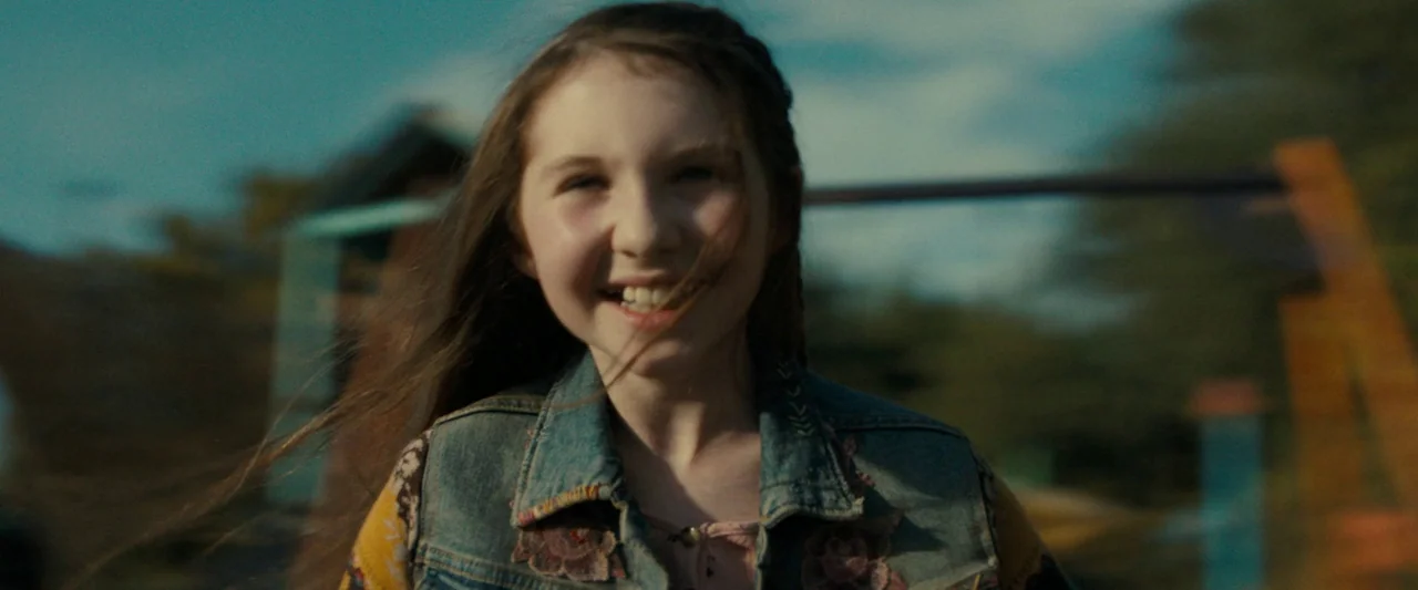Don’t Breathe 2 (2021) movie still: medium — An outdoor playground features out-of-focus structures against a backdrop of trees. A young girl smiles…