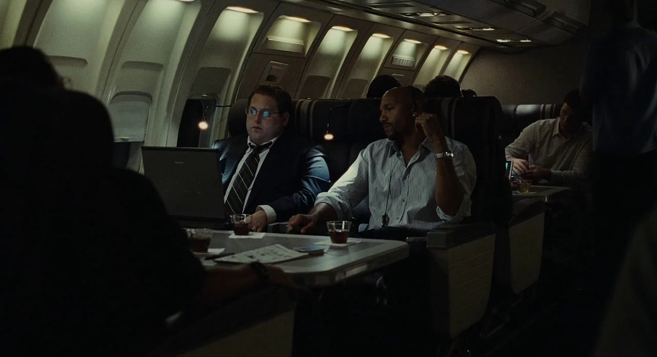 Moneyball (2011) movie still: medium — A dimly lit airplane cabin with dark upholstered seats and small individual reading lights. Two men sit at a…