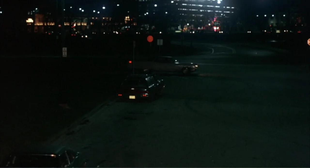 Cooley High (1975) movie still: wide — A dark, expansive parking lot at night is sparsely occupied by several parked cars. Distant city buildings…