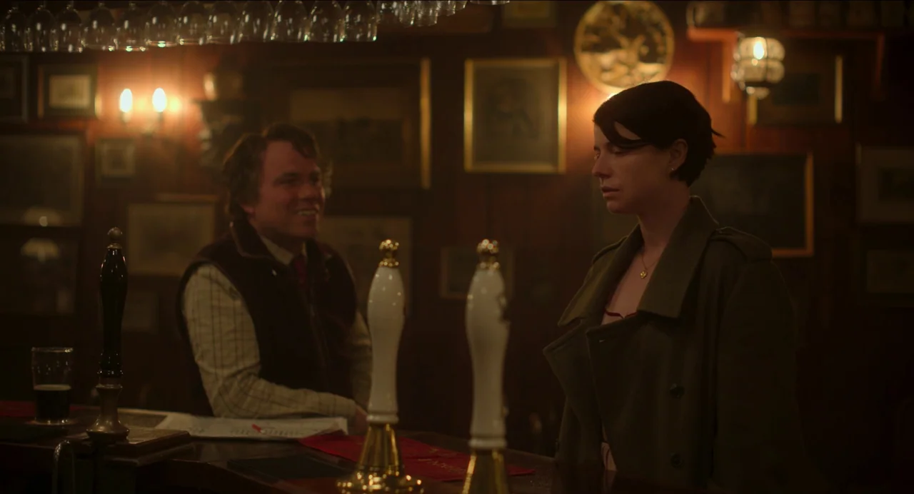 Men (2022) movie still: medium — A dimly lit pub interior features wooden walls covered in framed pictures and memorabilia. A woman in a dark coat…