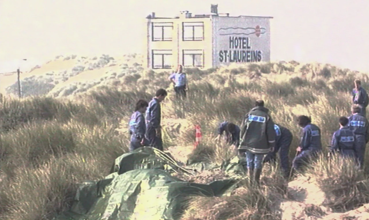 Maldoror (2024) movie still: wide — A group of people stand in sand dunes near a building labeled Hotel St-Laureins. The individuals examine a large…