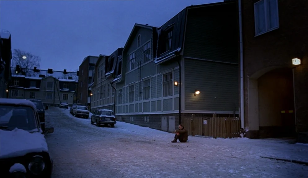 Night on Earth (1991) movie still: wide — A snowy street lined with wooden residential buildings under a deep blue twilight sky. A single man sits…