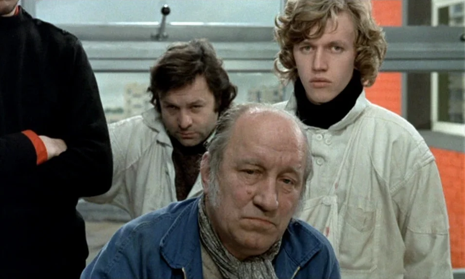 Tout Va Bien (1972) movie still: medium — Four men stand close together in an industrial workspace with a large window behind. They are positioned…