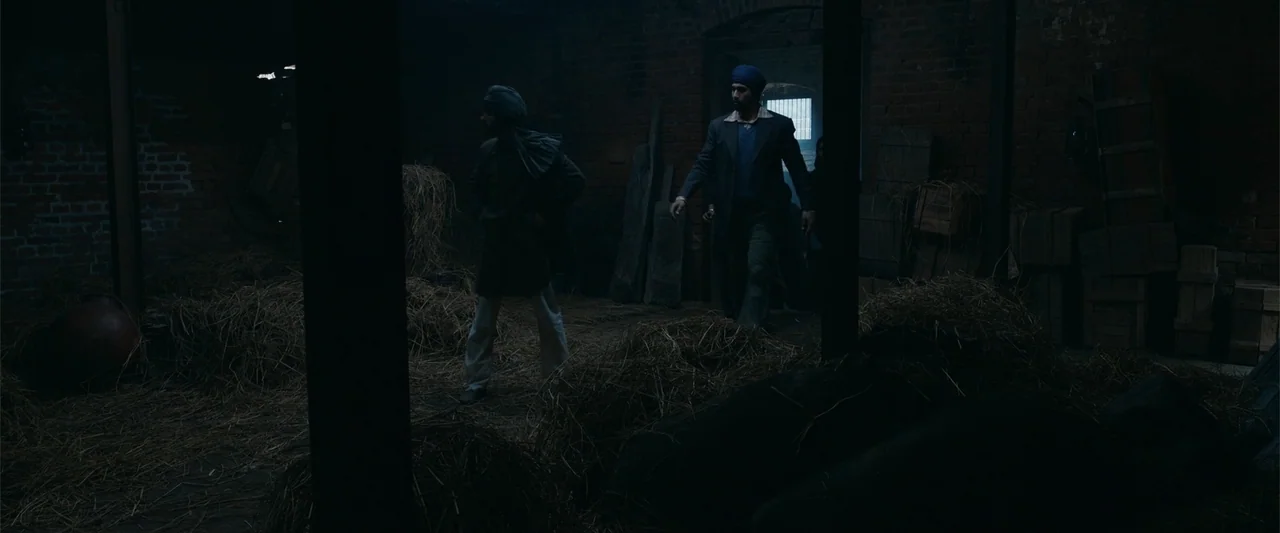 Sardar Udham (2021) movie still: wide — A dark brick building interior with stacks of hay and wooden crates. Two men walk across the space toward a…