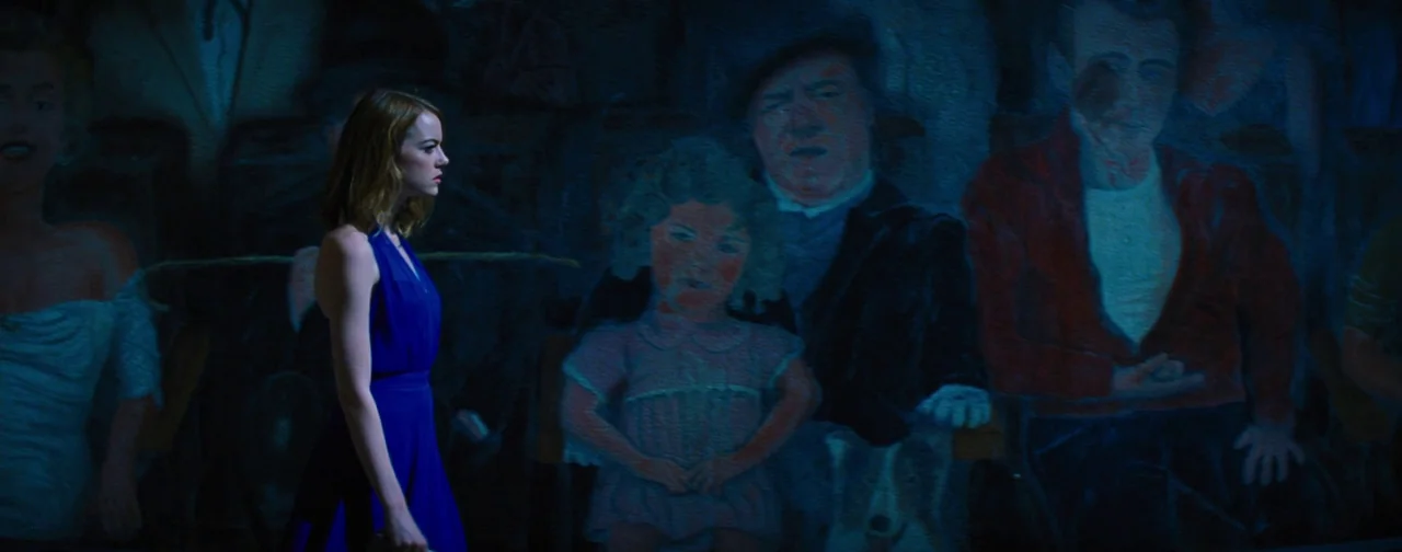 La La Land (2016) movie still: medium — A dimly lit indoor passage features a wall covered in a large painted mural. A young woman in a blue dress…