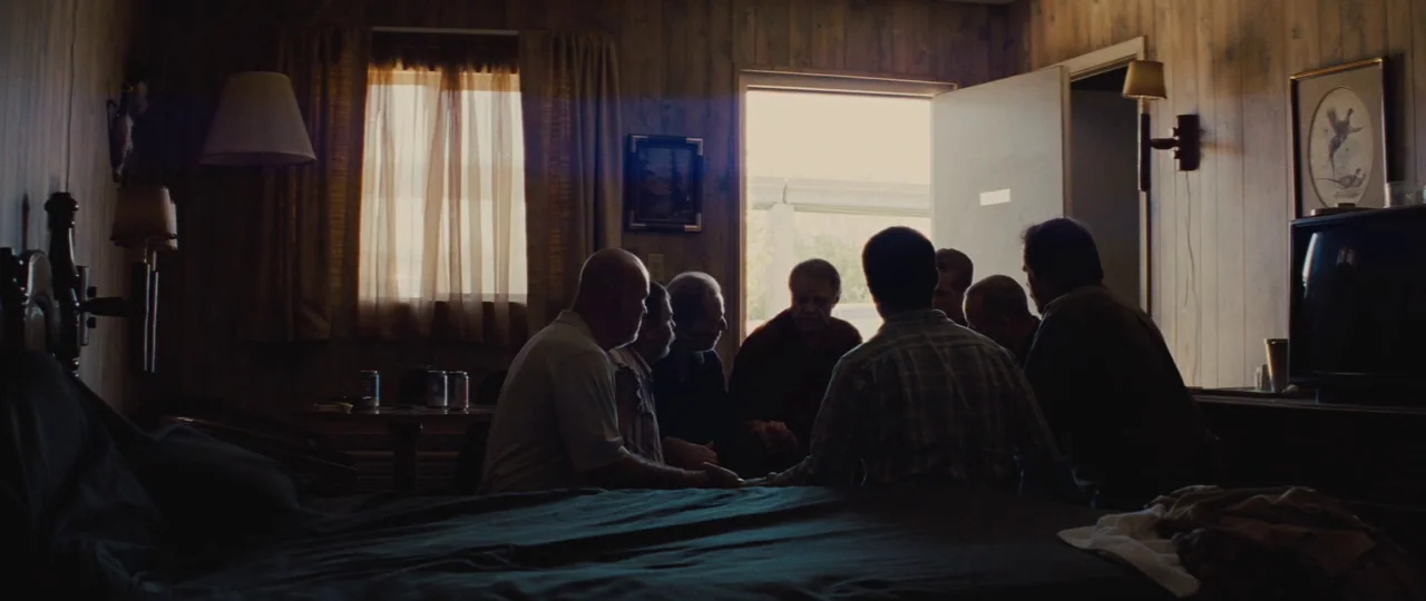 Mud (2012) movie still: wide — A dark motel room with wood-paneled walls features a bed occupied by several men. The group sits in a circle while…
