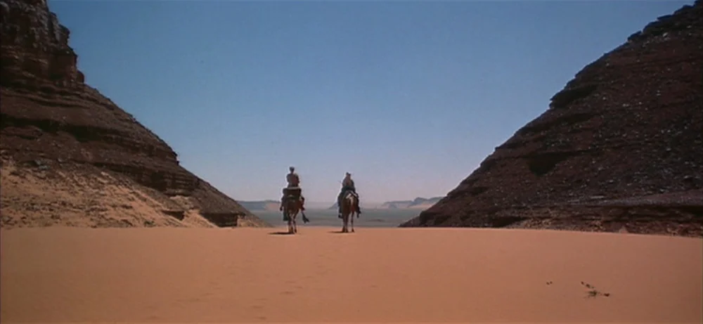 Lawrence Of Arabia movie still: wide — A vast sandy desert passage sits between two tall, craggy, deep-red rock formations. Two riders mounted on…