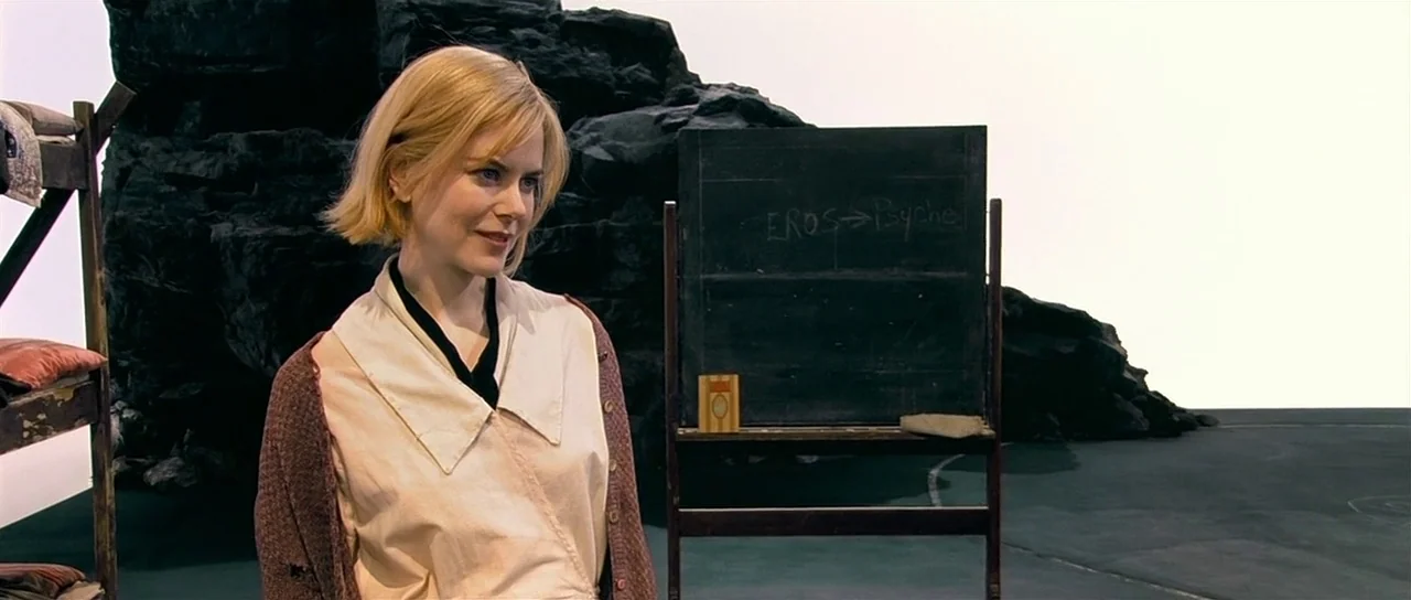 Dogville (2003) movie still: medium — A woman stands on a minimalist stage set consisting of rocky formations and a chalkboard. The chalkboard has…