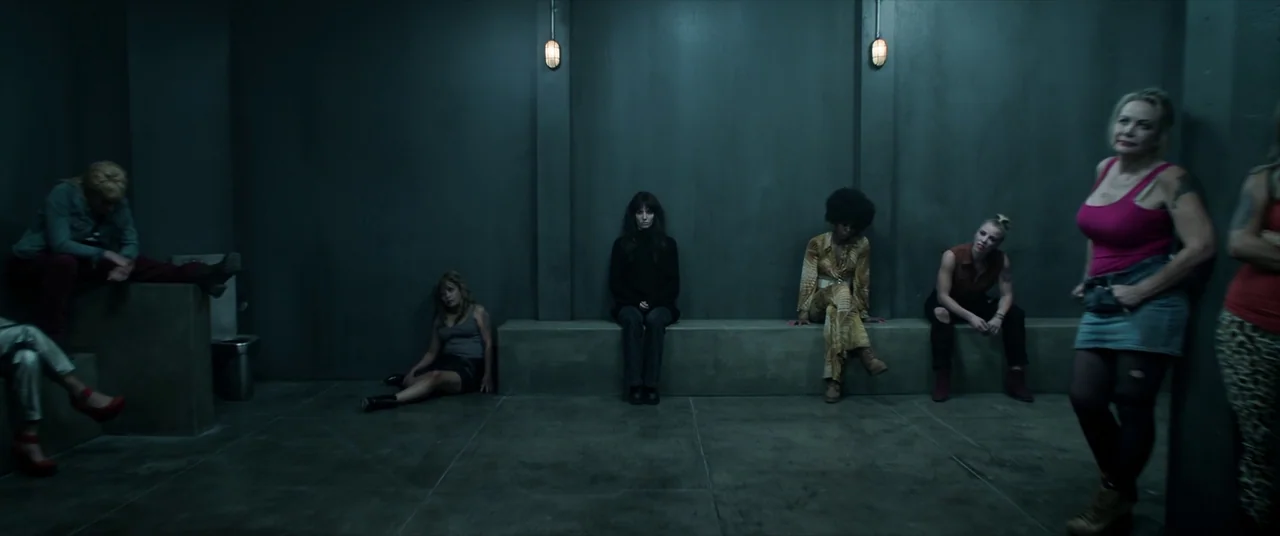 Malignant (2021) movie still: wide — A stark concrete holding cell features a long stone bench built into the wall. Seven diverse women in varied…
