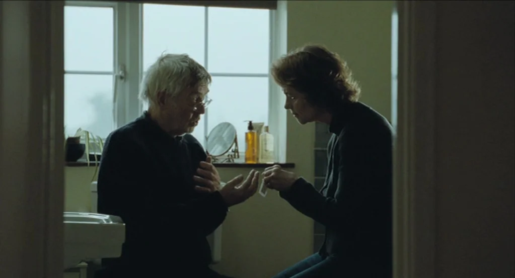 45 Years (2015) movie still: medium — A residential bathroom with a large window and a white pedestal sink. The man sits while the woman stands,…