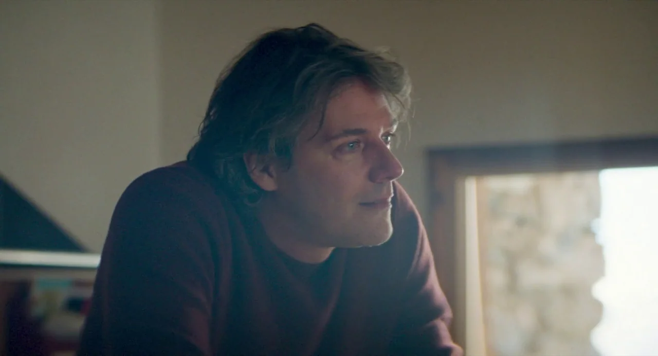Anatomy of a Fall (2023) movie still: medium — A dimly lit room features a man positioned near a bright window. The subject gazes towards the right…
