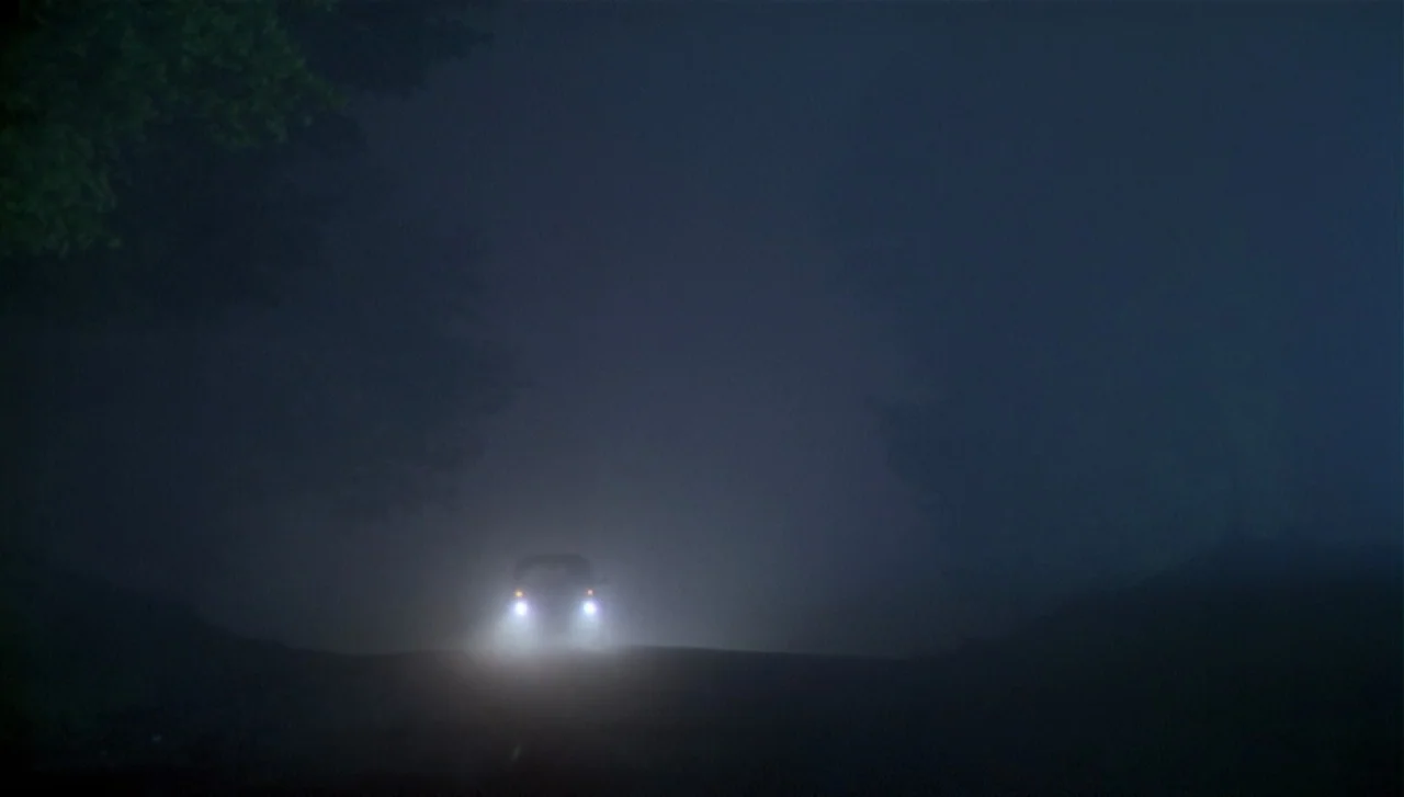 The Dead Zone (1983) movie still: establishing — A dark, unlit road is shrouded in thick fog at night. A lone vehicle approaches in the distance…