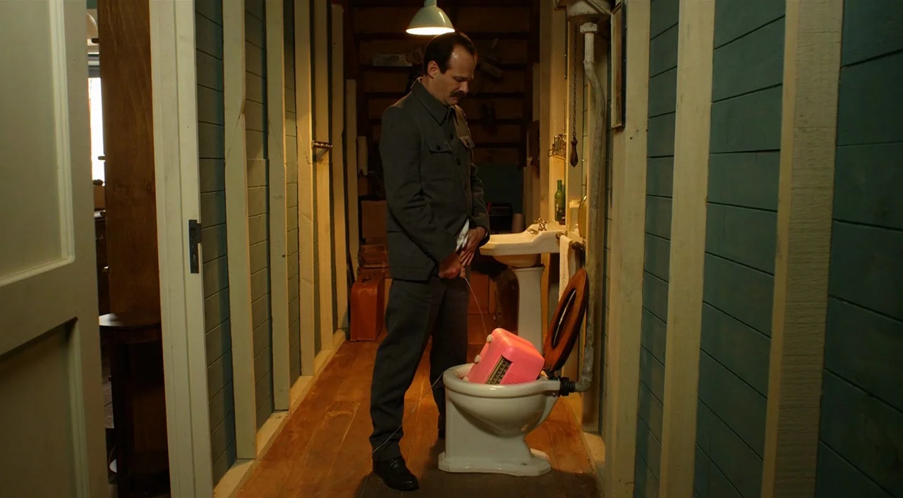 Dance of Reality (2013) movie still: fullbody — A cramped room with blue painted wooden walls and a simple ceramic toilet fixture. A man in a grey…