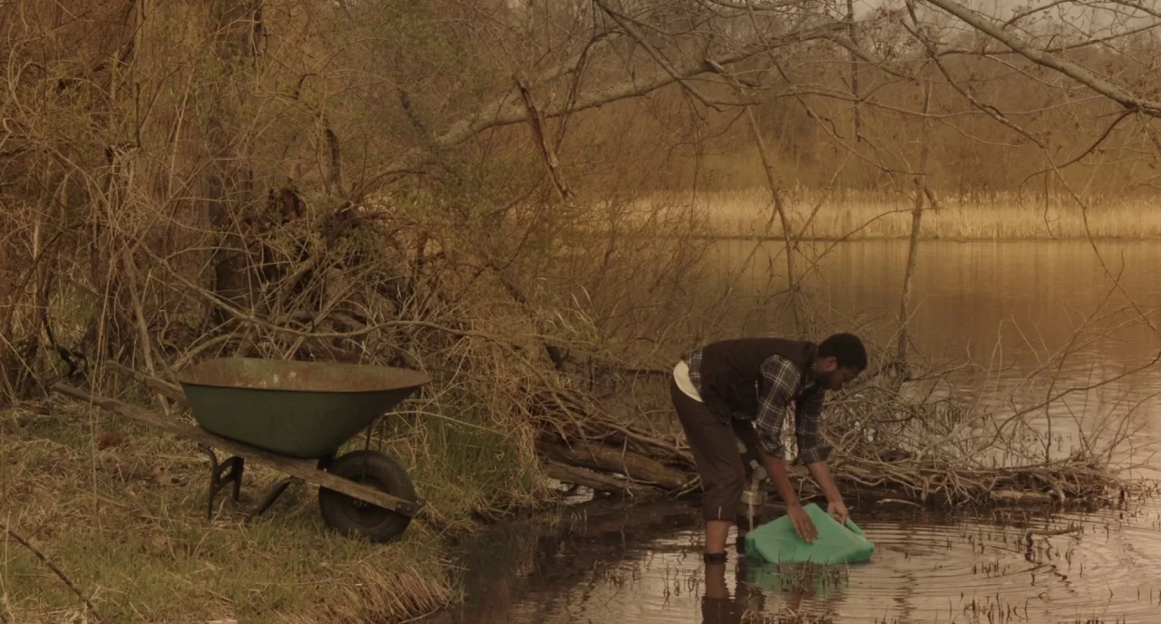 The Alchemist Cookbook (2016) movie still: wide — A rural lakeshore features dry brush, tangled tree roots, and a metal wheelbarrow nearby. A man…