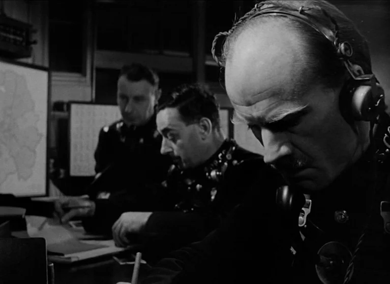 The Blue Lamp (1950) movie still: medium — A dimly lit room filled with men working at tables under low-key lighting. The subjects examine documents…