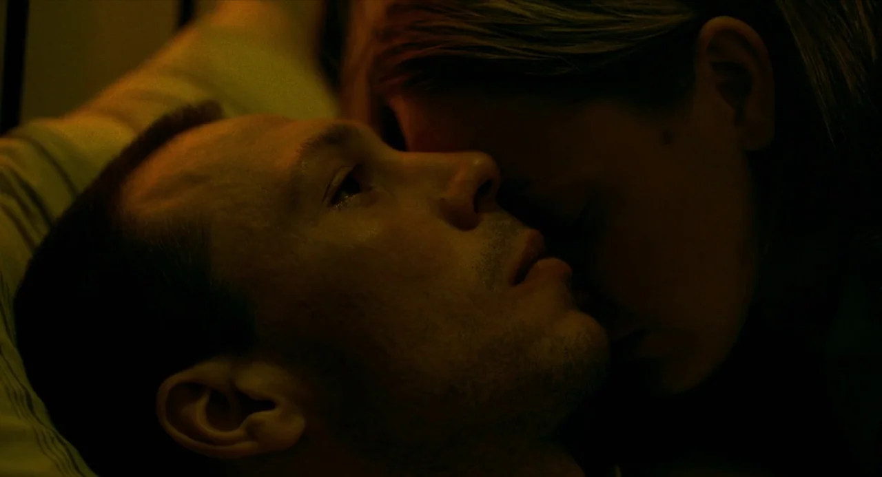 Beanpole (2019) movie still: closeup — Two people lie close together in a dark room under yellow light. The man faces up while the woman rests her…