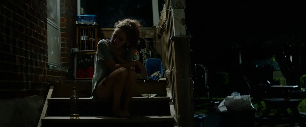 The Monster (2016) movie still: medium — A person sits on the wooden steps of a house porch at night. A liquor bottle and an ashtray are placed on…