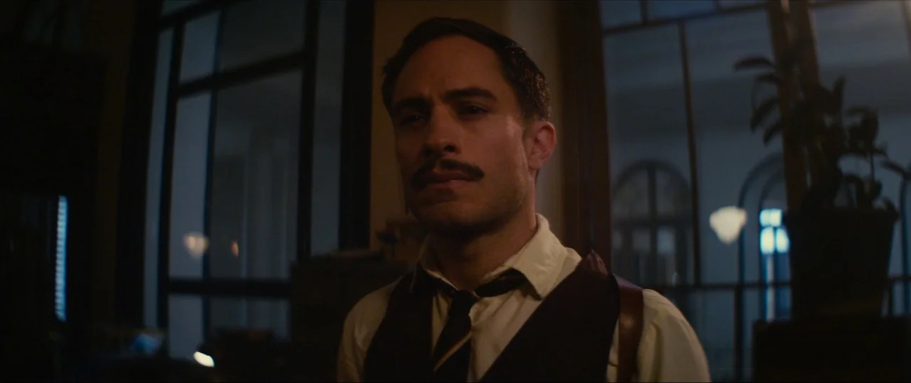 Neruda (2016) movie still: medium — A dimly lit room features large windows looking out into a dark evening sky. A man stands in the center of the…