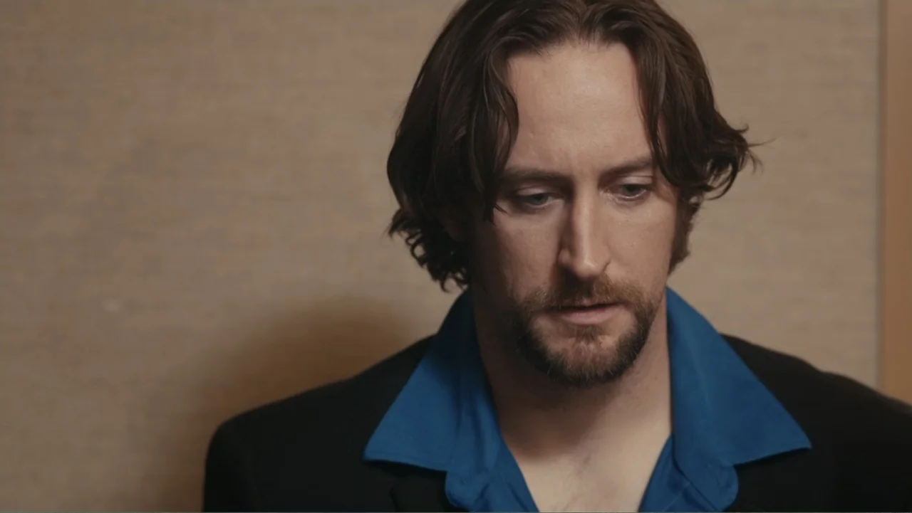 Toss It (2019) movie still: closeup — An interior space features a plain, textured beige wall as a background. A man with shoulder-length wavy hair…