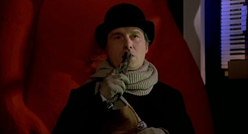 Malpertuis (1971) movie still: closeup — A man wearing a bowler hat sits before a large, textured red surface. He holds a small, possibly metallic,…