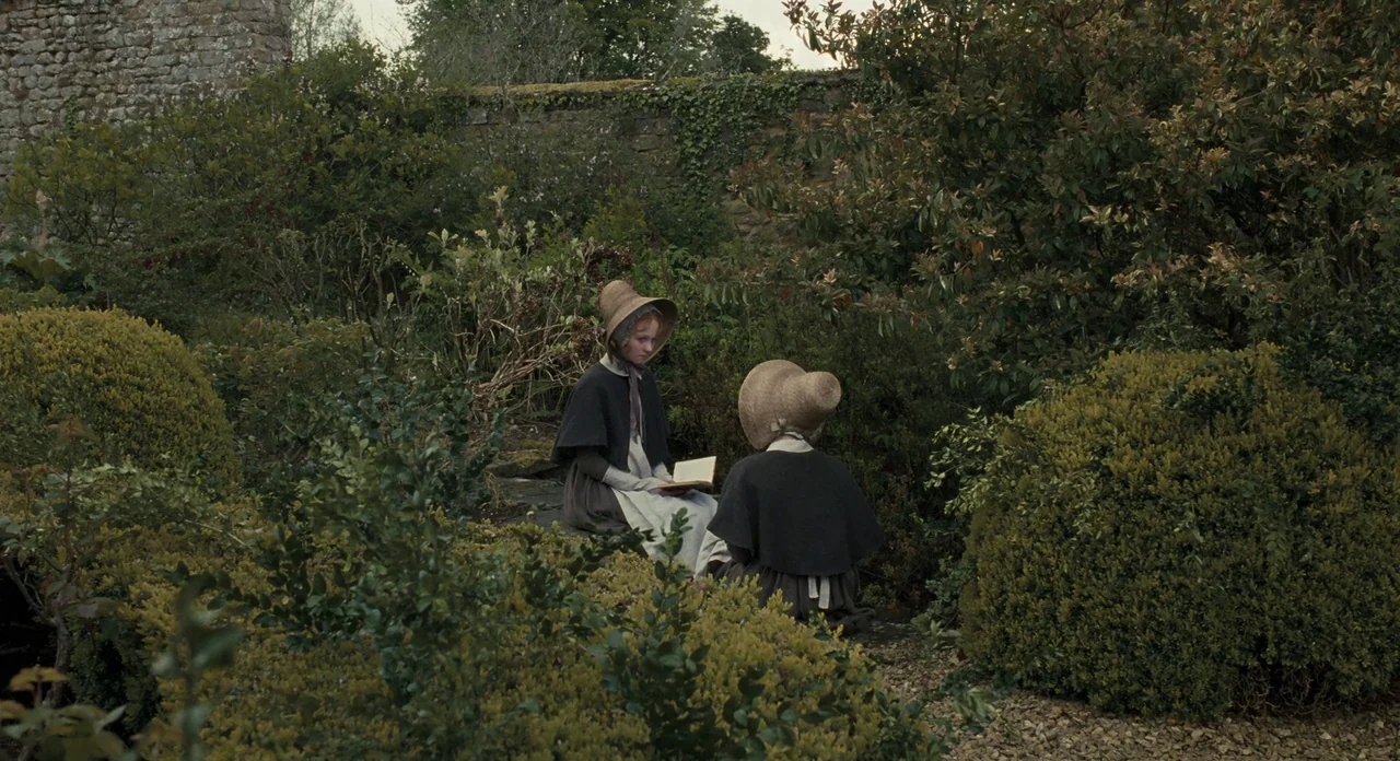 Jane Eyre (2011) movie still: wide — A dense outdoor stone-walled garden is filled with thick shrubs and green foliage. Two women in period clothing…