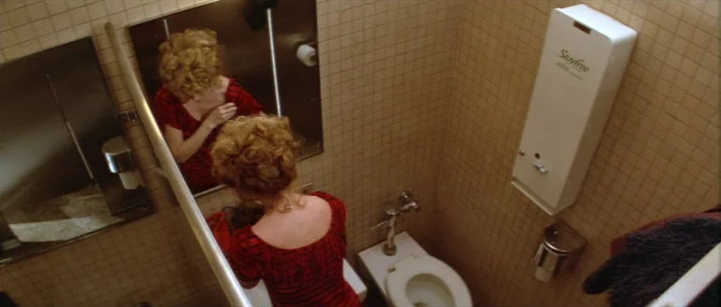 Blow Out (1981) movie still: medium — A cramped tiled public restroom features tan square tiles on the walls. A woman stands in front of a…