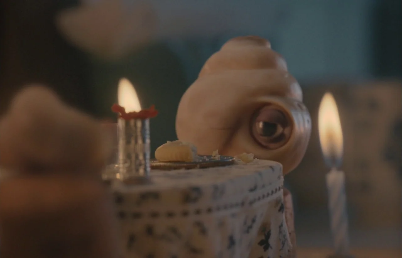 Marcel the Shell with Shoes On (2021) movie still: closeup — A small, rounded creature sits at a table covered in a patterned, embroidered cloth.…