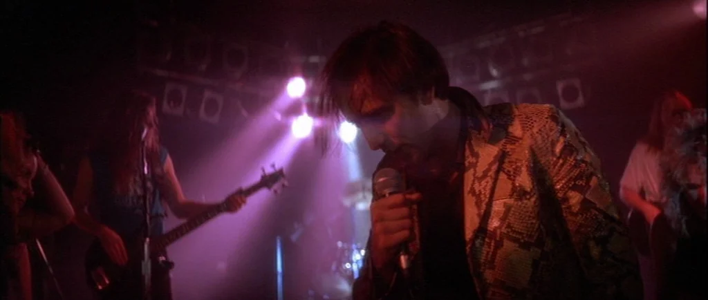 Wild At Heart (1990) movie still: medium — A dimly lit musical performance venue features intense stage lights overhead. A male singer in a…