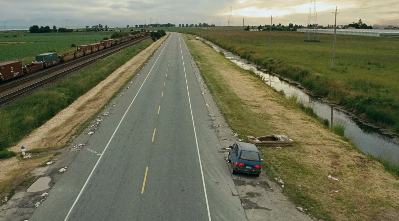 Juno (2007) movie still: establishing — A long straight asphalt road cuts through a rural landscape with rail tracks nearby. A small blue car is…