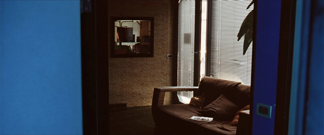 GOMORRAH (2023) movie still: wide — A modern interior room features a brown sofa placed against a textured stone wall. Light streams through window…