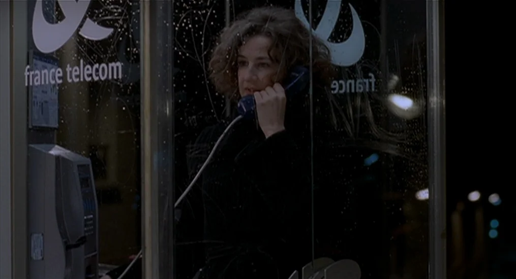 Vendredi Soir (2002) movie still: medium — A nighttime exterior featuring a woman inside a glass-walled public phone booth. Rain droplets coat the…