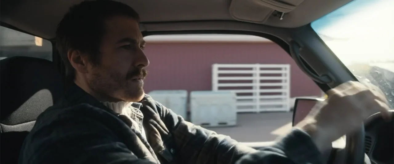 A Small Fortune (2021) movie still: medium — A man drives a truck during the day with a red building visible outside. The driver steers the vehicle…