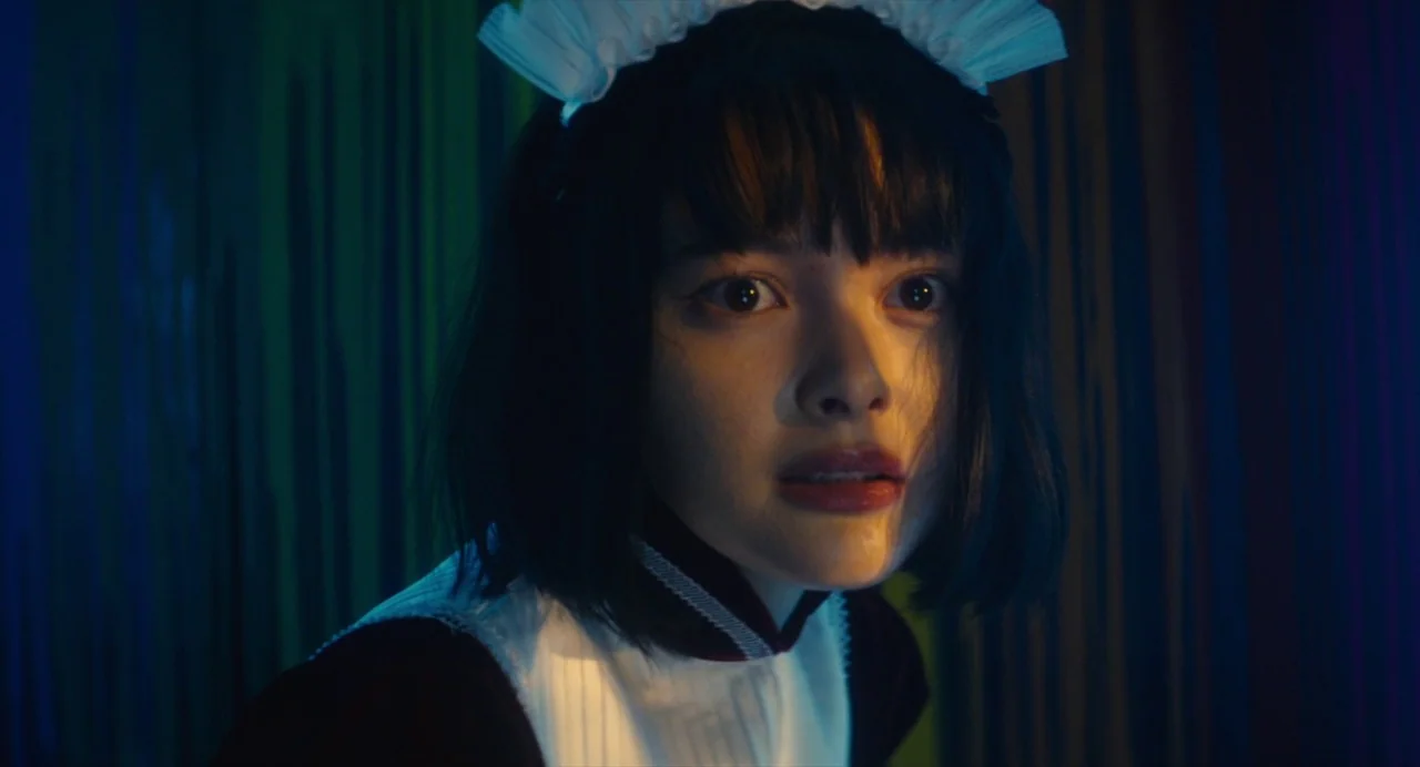 Diner (2019) movie still: closeup — A dark space features a woman in a maid uniform with neon-tinted background curtains. The woman looks directly…