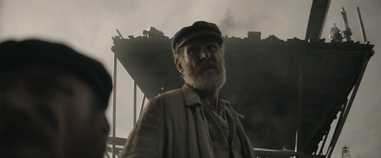 Eiffel (2021) movie still: medium — A construction site features exposed wooden scaffolding and metal beams under a cloudy sky. An older man with a…