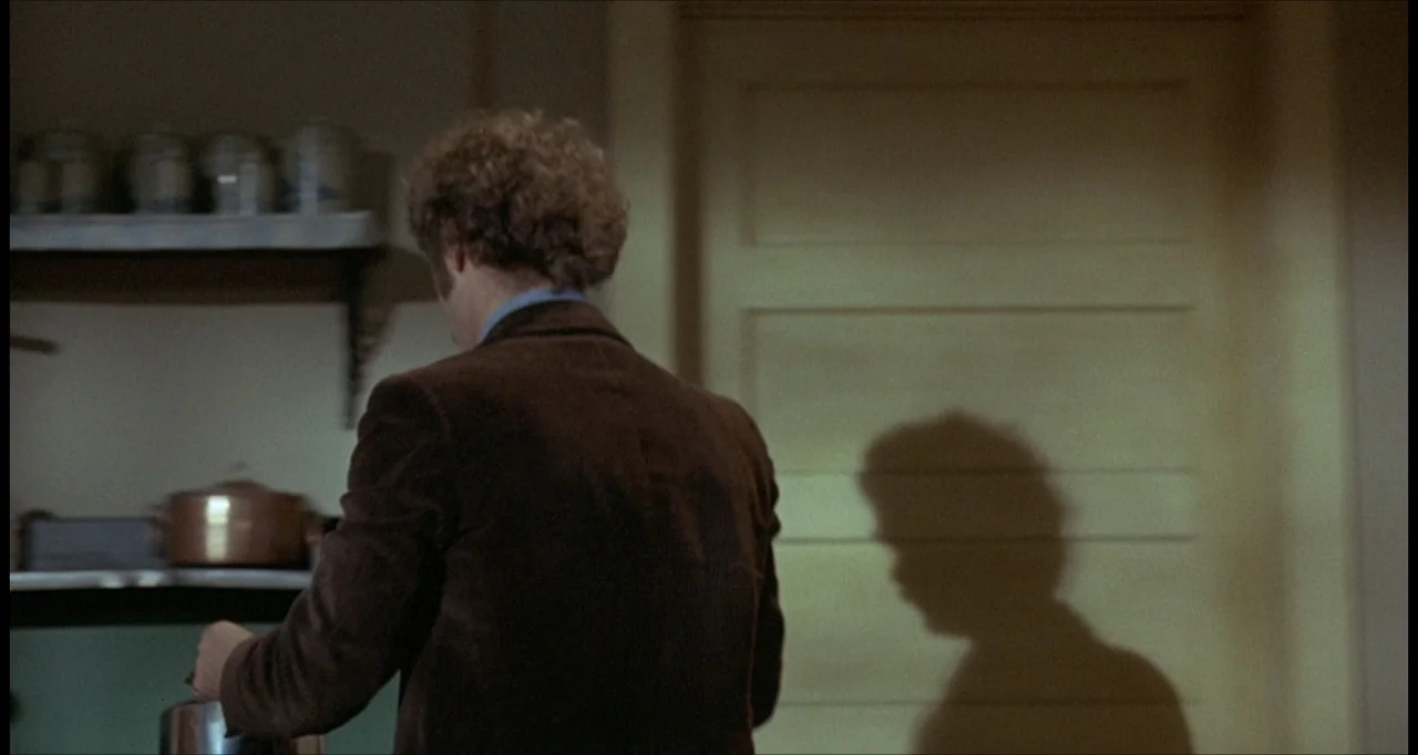 The Dunwich Horror (1970) movie still: medium — A dimly lit room features a plain wall and kitchen shelves with containers. A man in a brown jacket…