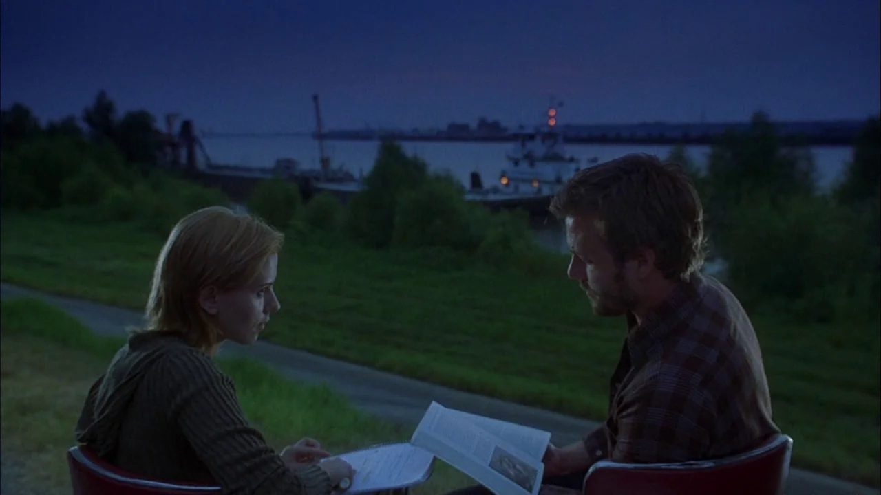 Love Song For Bobby Long (2004) movie still: medium — A man and woman sit in red chairs on a grassy riverbank at twilight. They read from a book…
