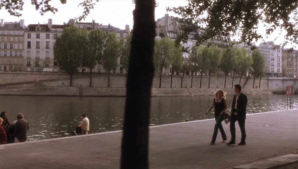 Before Sunset (2004) movie still: wide — A stone embankment lines the Seine river in Paris with historic buildings in the background. A man and…