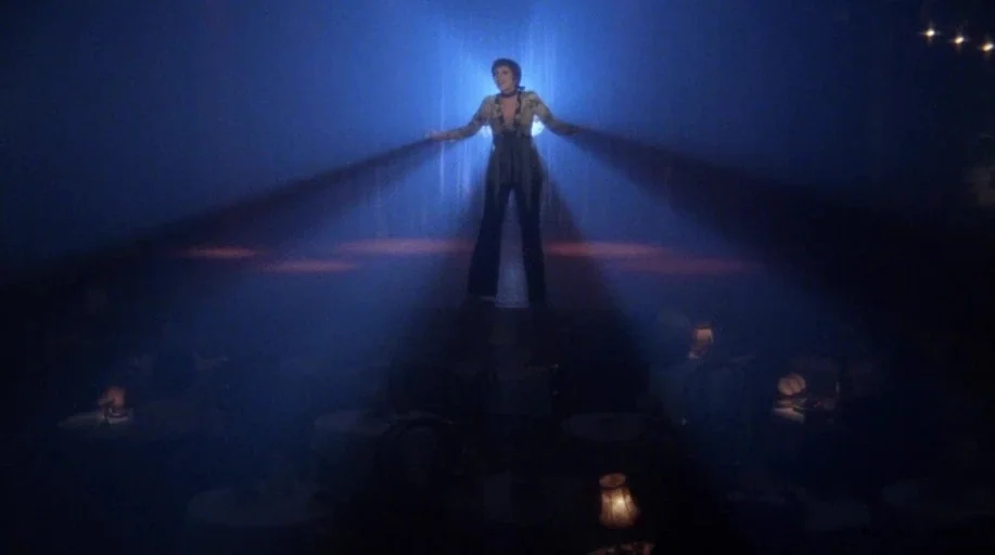 Cabaret (1972) movie still: fullbody — A dimly lit nightclub with tables scattered across a dark, smoky floor space. A woman stands centered on a…