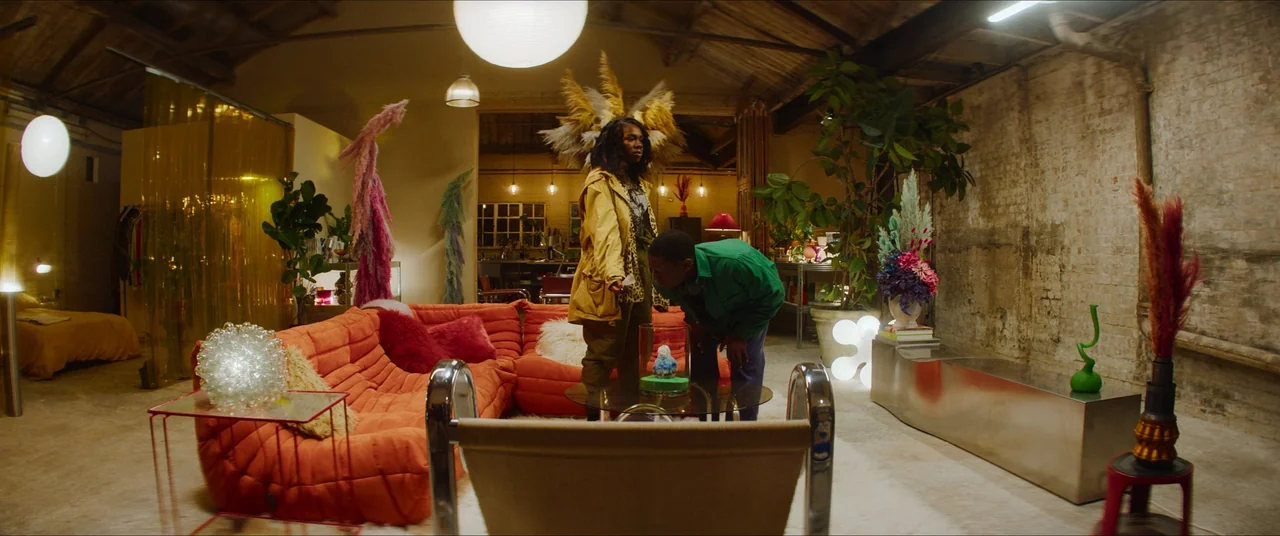 Rye Lane (2023) movie still: wide — A spacious industrial loft features a large orange sofa, decorative dried grasses, and exposed brick walls. A…