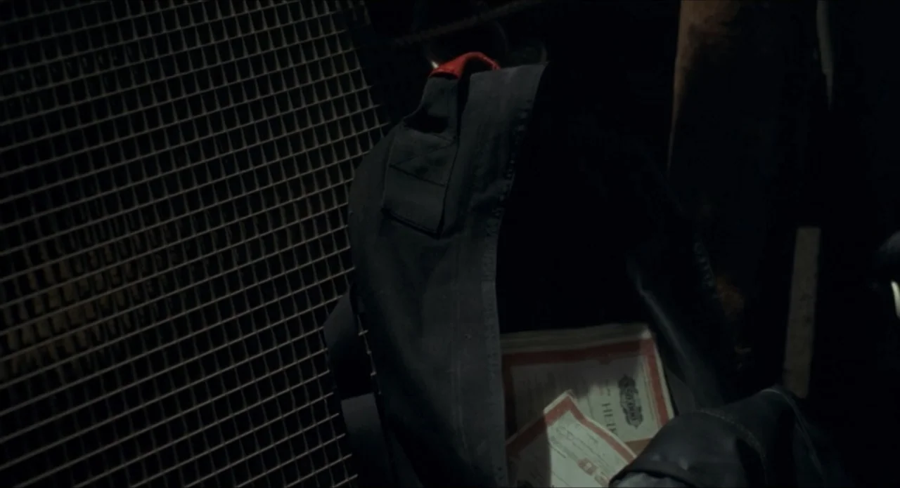Foolproof (2003) movie still: closeup — A dark, cramped space features a black backpack resting against a wire metal grate. Several stacks of…