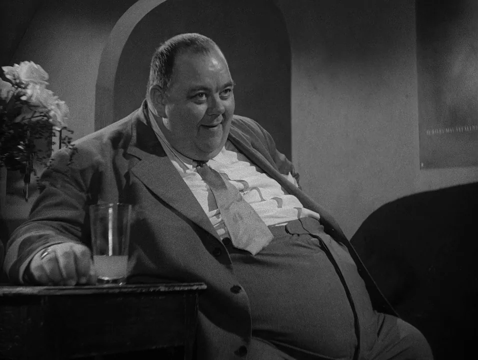 Dreams (1955) movie still: medium — A dimly lit room features a heavy-set man seated at a small wooden table. A glass of liquid sits on the table…