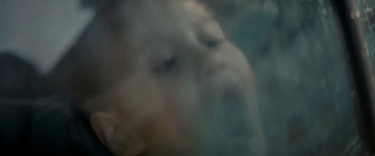 Ali & Ava (2021) movie still: closeup — A dark, out-of-focus view of a child's face pressed against a glass pane. The child's mouth is partially…