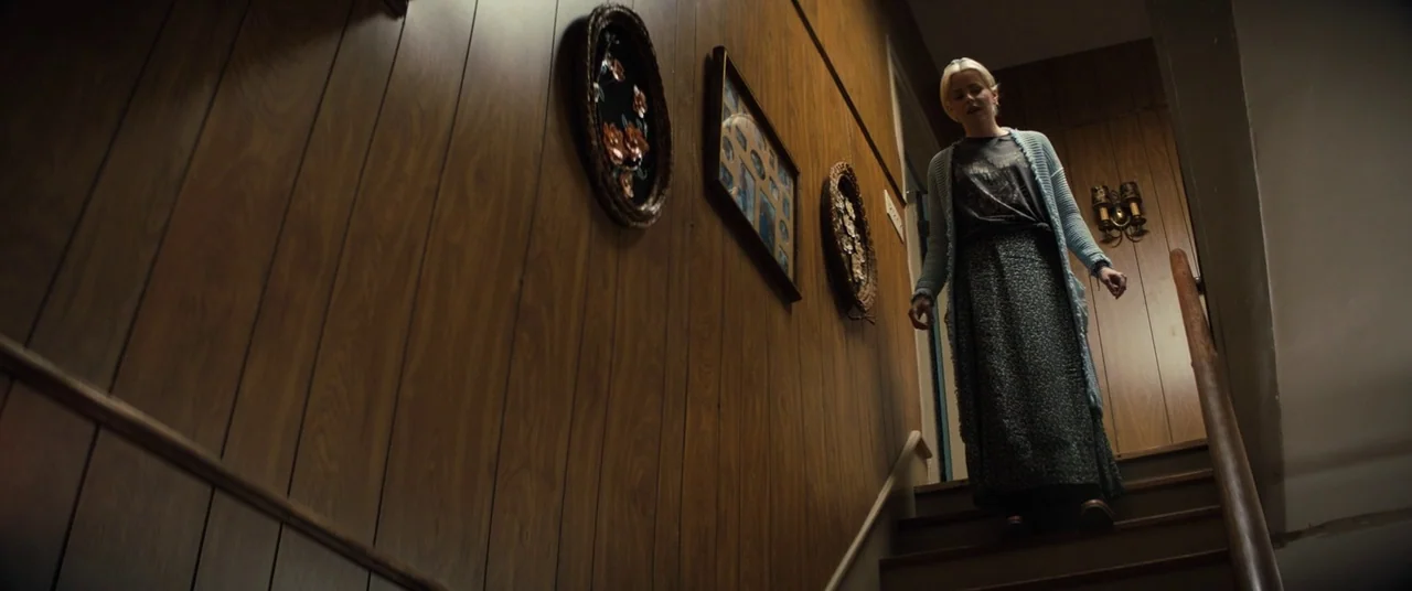 Brightburn (2019) movie still: fullbody — A residential staircase features brown wood-paneled walls decorated with framed pictures and a sconce. A…