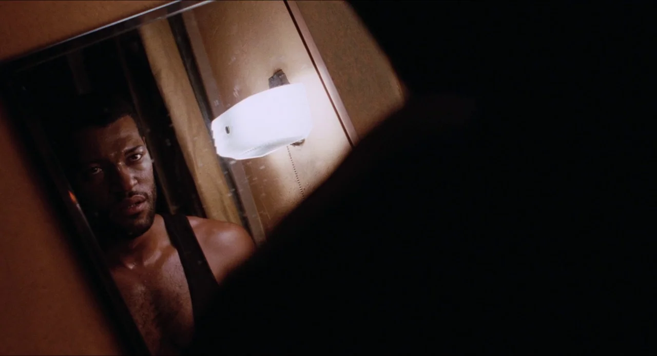 Deep Cover (1992) movie still: closeup — A dimly lit room with a man standing in front of a glass surface. The subject is gazing into the mirror…