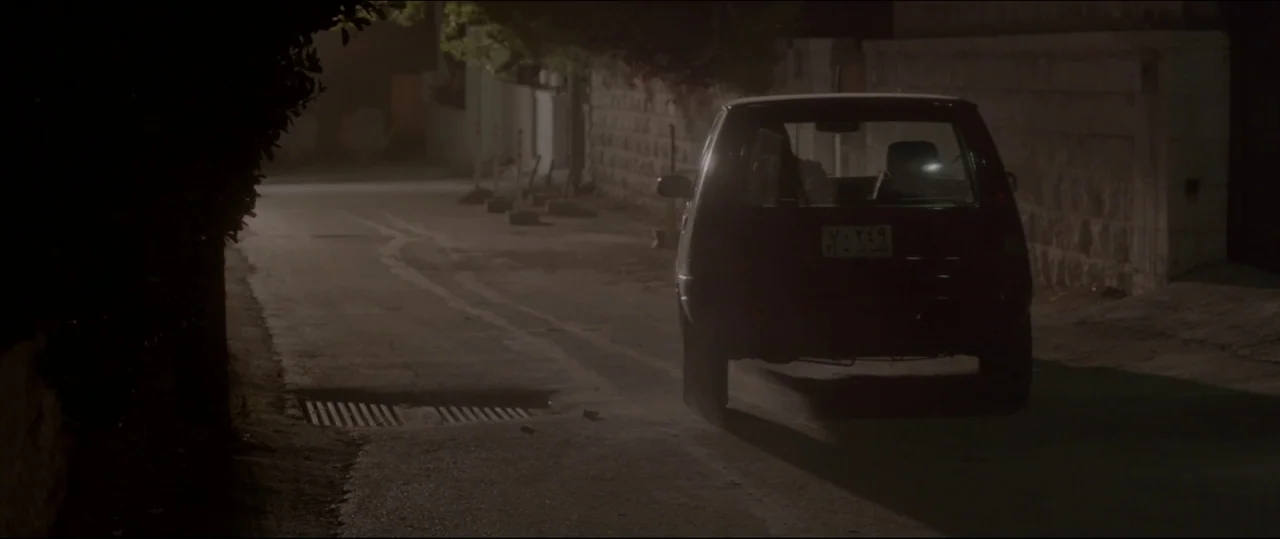 Under The Shadow (2016) movie still: wide — A narrow suburban residential road at night is bordered by a stone wall. A dark-colored hatchback car is…