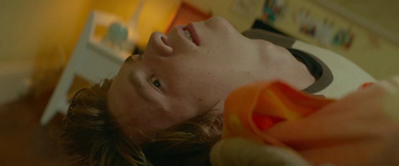 Me and Earl and the Dying Girl (2015) movie still: closeup — A teenage boy lies on a surface looking upward in a dimly lit room. A blurred chair and…