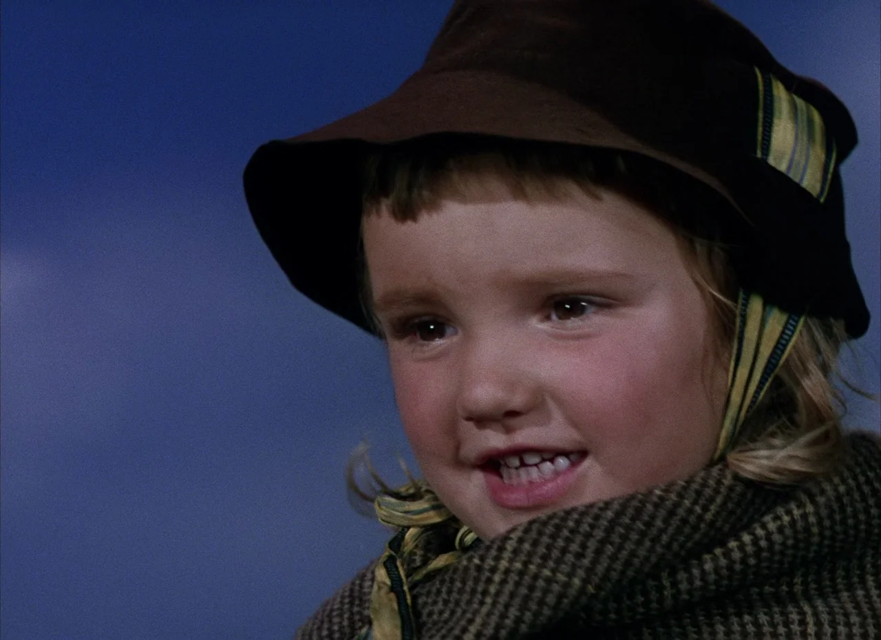 Shane (1953) movie still: closeup — A young girl is positioned against a solid, dark blue background. She is wearing a brown hat and a thick wool…
